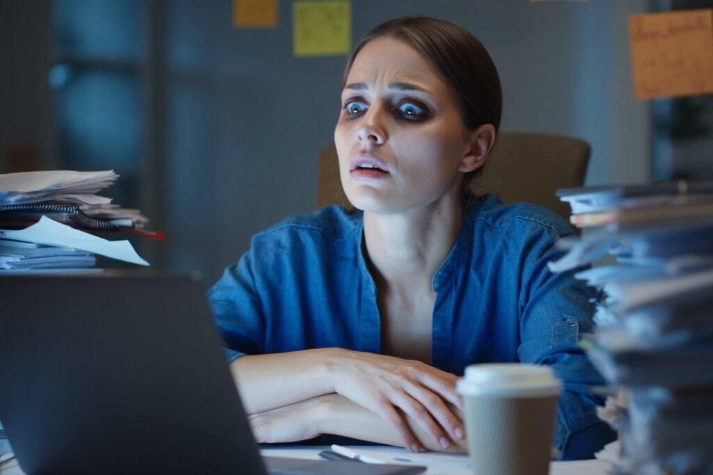 a woman in her 30s sitting at a cluttered desk late at night, perfectly capturing the 'wired but tired' state of chronic stress and high cortisol
